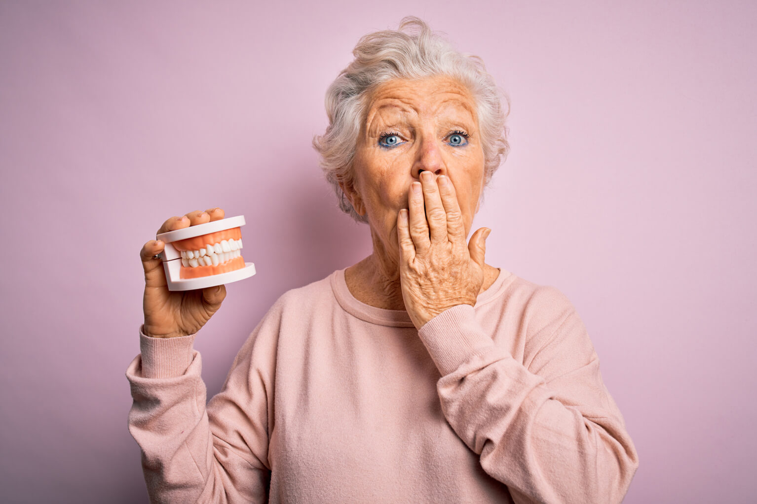 Senior beautiful grey-haired woman holding plastic denture teeth over pink background